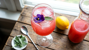 High-angle shot of a sparkling pink guava mocktail served in a wine glass over ice, garnished with a purple edible flower and a fresh sage leaf. A carafe of pink pre-batched base, a whole lemon, a bowl of sage leaves, and a bar spoon sit on a rustic wooden table next to the drink