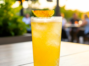 A chilled golden sparkling cocktail in a tall highball glass, topped with a lime wedge dusted with chili, served on an outdoor table at sunset with warm bokeh lights in the background.