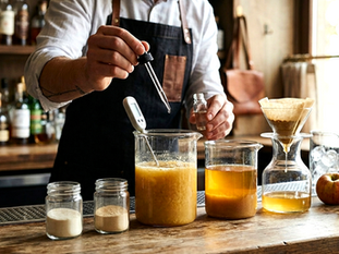 A bartender using a dropper to add enzymes to a beaker of fruit pulp, with jars of powder and a clarified apple cocktail on a wooden bar.