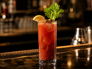 An elegant Bloody Mary in a tall highball glass on a dark wood bar, garnished with a single short celery stalk and a thin lemon wheel.