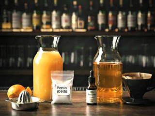 A professional bar counter displaying the juice clarification process. On the left, a carafe of cloudy orange juice sits next to a citrus reamer. Center stage features a bag of Pectin (E440) powder and a bottle of Pectinex enzyme. On the right, a carafe contains the final clarified, transparent liquid next to a coffee filter setup, illustrating the before-and-after results of enzymatic clarification