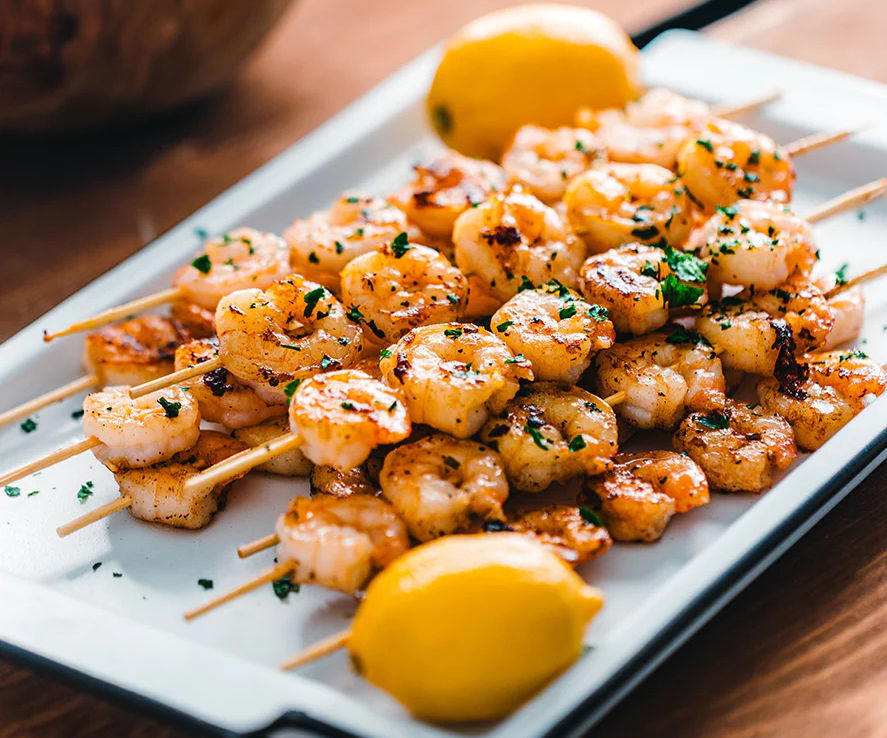 Grilled shrimp skewers garnished with herbs on a white platter, accompanied by lemons, on a wooden table. Bright and appetizing.