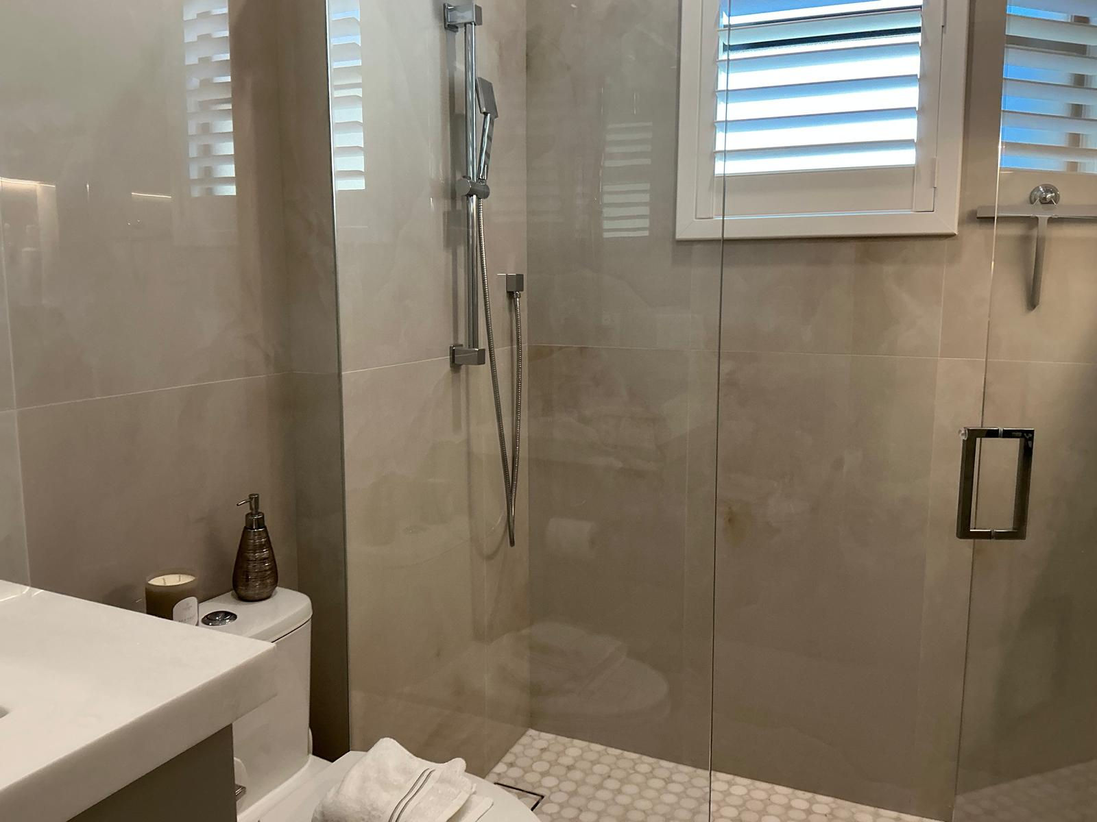 Modern bathroom with tiled shower, glass enclosure, white plantation shutters.