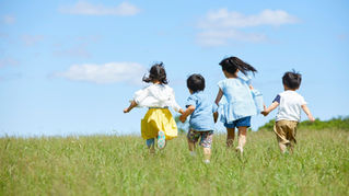 Four children run in a grassy field under a bright blue sky. They wear colourful clothes, creating a joyful and playful atmosphere.