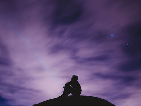 A lone figure gazes up at a starry night sky, silhouetted against swirling, illuminated clouds.