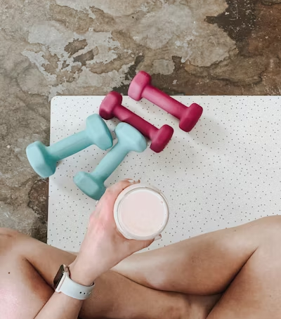 a person sitting cross-legged on a mat holding a protein shake. There are 2 sets of light dumbells placed on the ground by their legs.