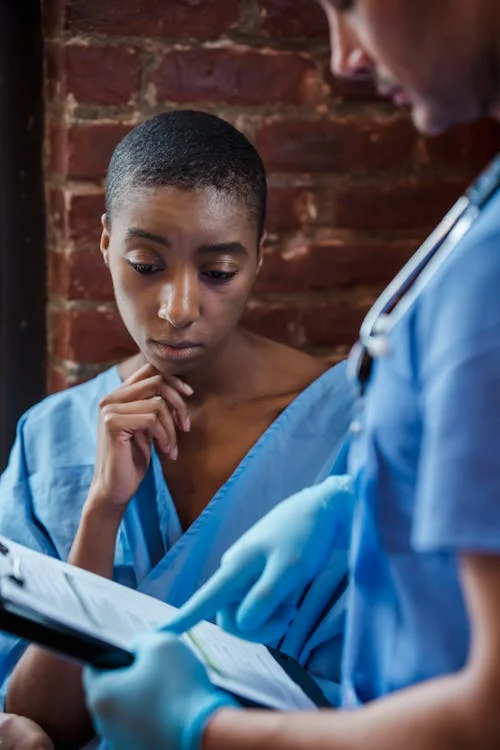 A Black woman in a hospital gown looking at a clipboard held by a male doctor