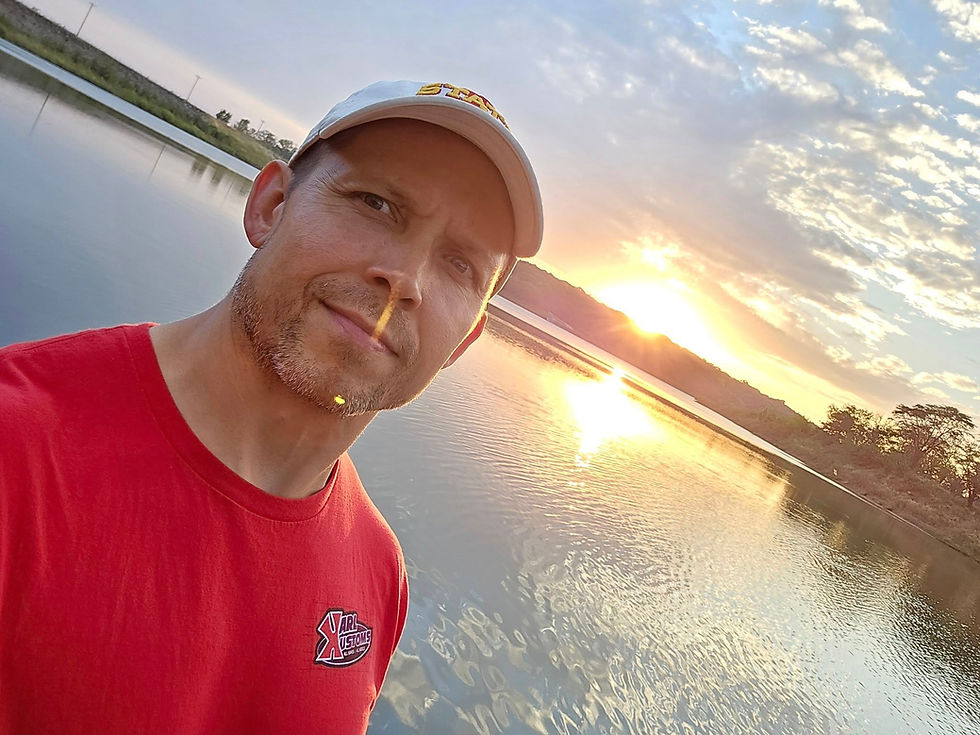 Man in red shirt and white cap taking a selfie by a serene lake at sunset. The sky is partly cloudy with warm, orange hues.