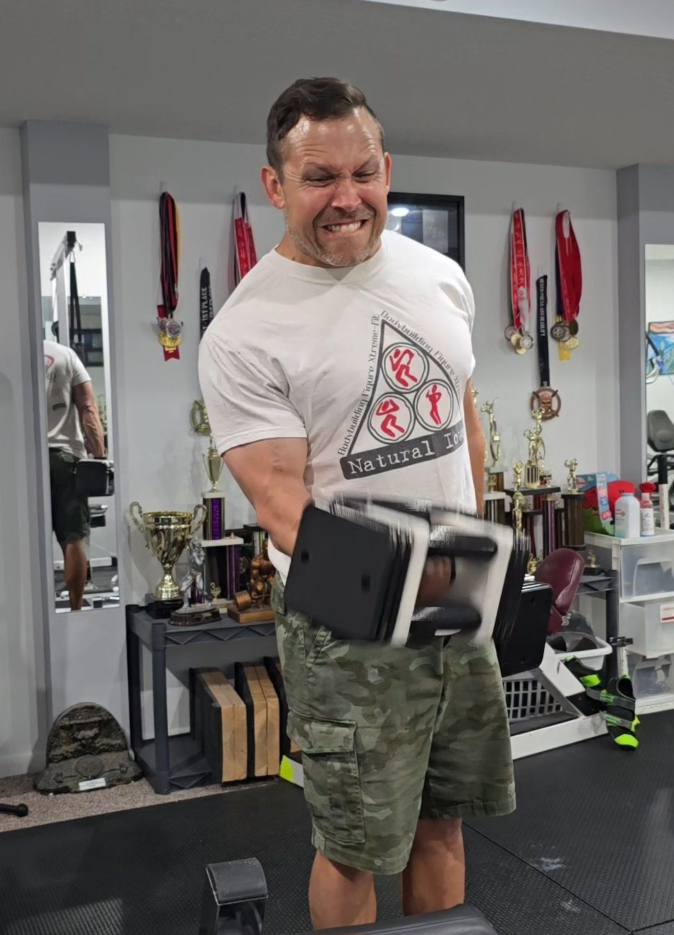 Man lifting dumbbell, grimacing in effort, wearing a white shirt and camo shorts. Trophies and medals line the gym's wall behind him.