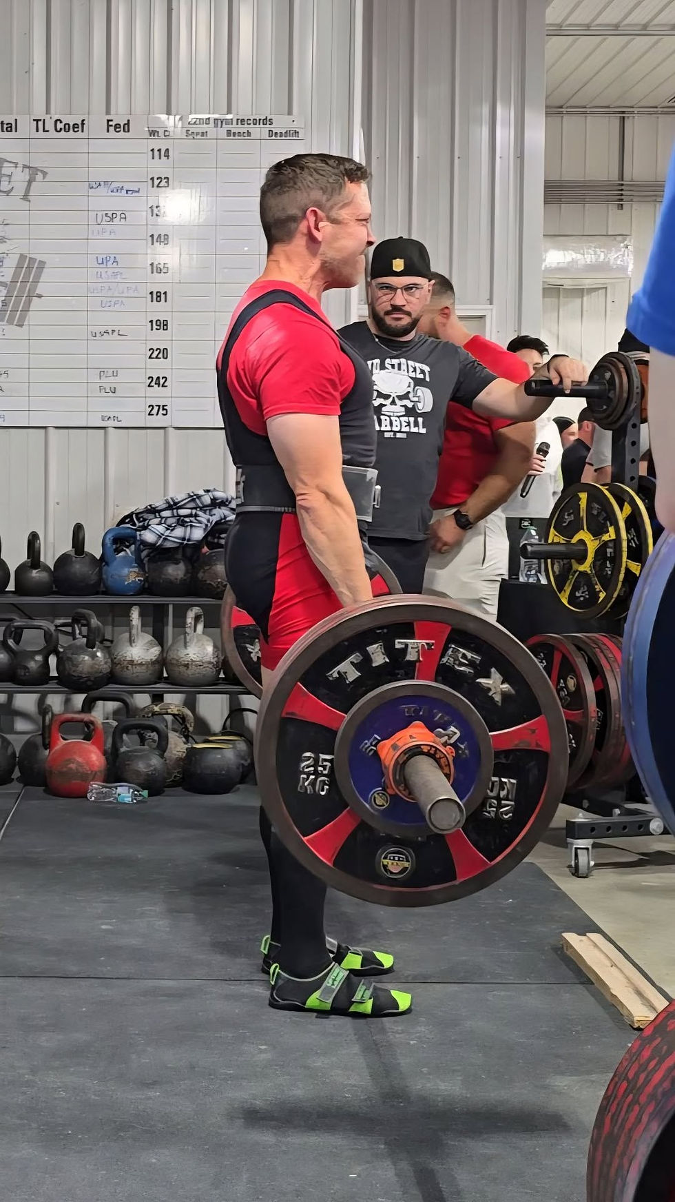 Man in red lifts barbell in gym, others watch. Kettlebells and weights in background. Chart on wall. Focused effort.