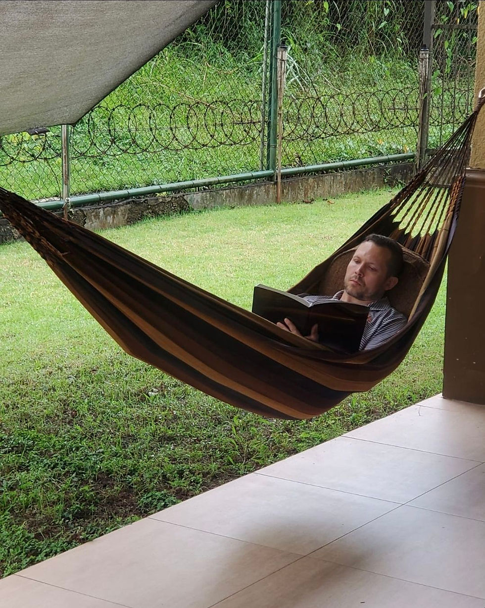 Man relaxing in a brown hammock, reading a book outdoors. Green grass and metal fence in the background. Calm, peaceful setting.