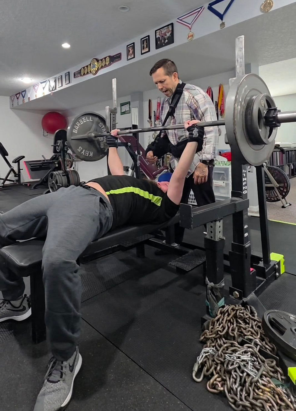 Man bench pressing weights, assisted by another in a gym room with medals on walls. Chains and weight plates visible, focusing on strength.