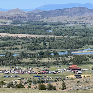 The Big Thing festival with many cars, tents, and a red pagoda-style building beside a river, backed by hills and blue mountains.