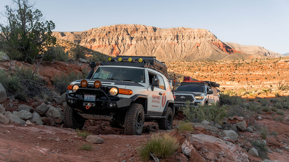 Two off-road vehicles navigate a rocky trail in a canyon, with a rugged landscape and clear sky. Text on lead vehicle reads "Four Points Adventures."