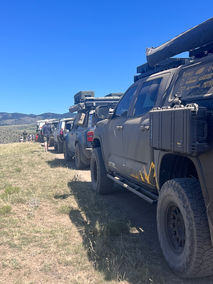 Overland rigs in a lineup at The Big Thing on a dirt trail with mountains in the background and blue skies.