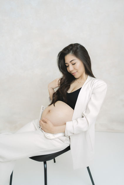 An expecting mother wearing a suit sits on a black chair smiling at her bump.