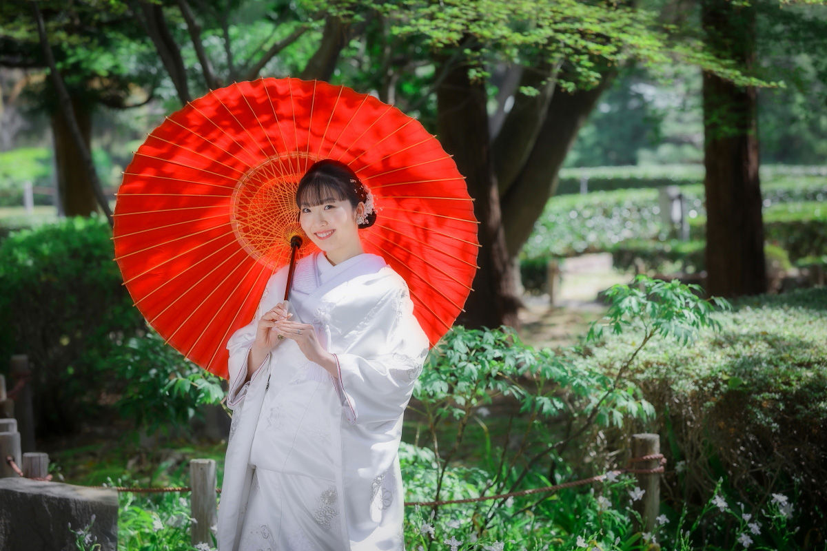 赤い和傘を手にした新婦が緑豊かな庭園で微笑む、名古屋での和装前撮りの一コマ