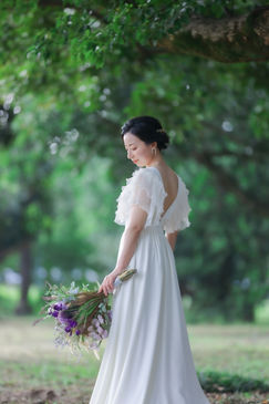 名古屋の庄内緑地公園で自然に囲まれた穏やかな光の中、新婦が後ろ向きに立ちながら振り返る優雅な姿と手に持ったナチュラルなブーケが印象的な前撮りシーン