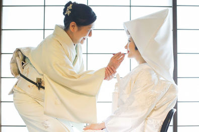 白無垢姿の新婦に向けて、母親が優しく化粧を施す、愛情あふれる室内撮影の一コマ