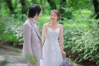 緑が生い茂る道を歩く新郎と新婦、名古屋の庄内緑地公園でのフォトウェディング。新郎はベージュのスーツに薄紫のネクタイを合わせ、新婦は繊細なレースが施された白いウェディングドレスを着用。お互いの目を見て微笑む、幸せな一瞬