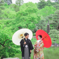 名古屋の日本庭園を背景に、赤と白の和傘を手にした和装の新郎新婦が向かい合う前撮りシーン