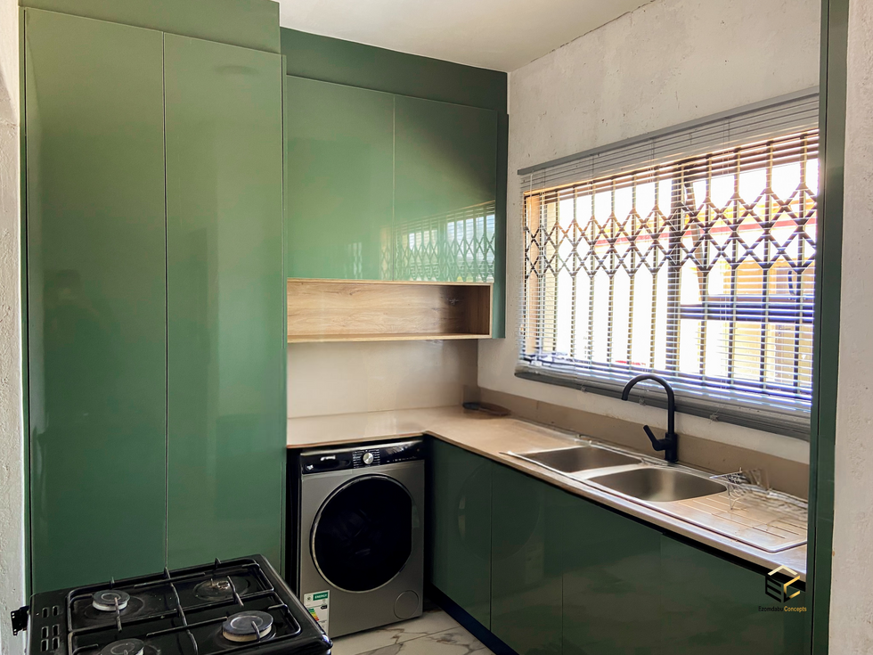 Green high-gloss kitchen cabinets with flush handles, integrated washing machine, double sink, black tap, and wooden floating shelf.