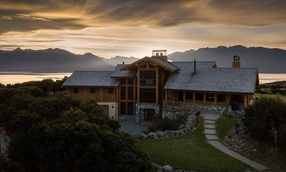 Fiordland Lodge at sunset, framed by mountains and a lake. Dramatic clouds fill the sky, casting warm hues over the serene landscape.