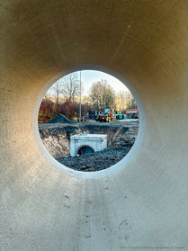 Blick durch ein Betonrohr auf eine Tiefbaustelle mit Schachtbauwerk und Baumaschinen