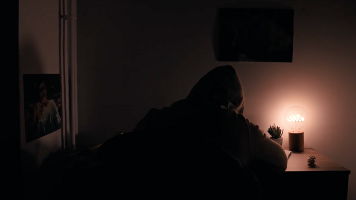 Figure encapuchonnée penchée sur un bureau, éclairée par une lampe unique dans l'obscurité.