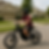 A person in a burgundy shirt rides a black dirt bike on a suburban street. The background shows beige houses and green lawns.