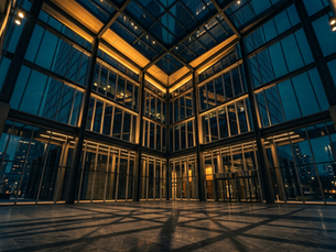 Low-angle view of a modern glass office building exterior at night, with warm gold lighting along the structural beams, geometric shadow patterns on the dark stone floor, and a city skyline visible through the glass panels in the background.