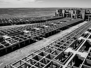 Aerial view of a vast brutalist concrete grid structure in a barren desert landscape, three distinct sections of unfinished frameworks converging toward a monolithic block of dense concrete forms on the horizon, harsh overhead light casting deep shadows across the geometric surface, black and white.