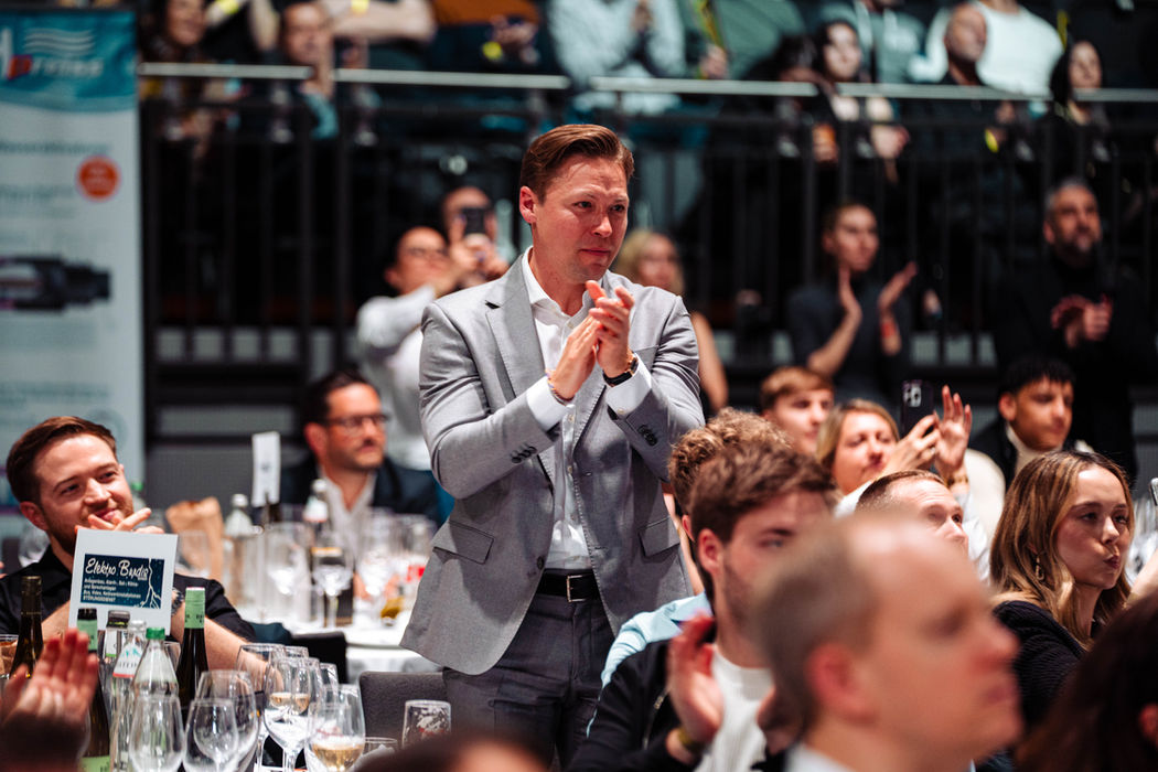 Ein Mann im grauen Sakko applaudiert stehend, umgeben von einem vollbesetzten Publikum – begeisterte Reaktionen bei der Bounce Fight Night.