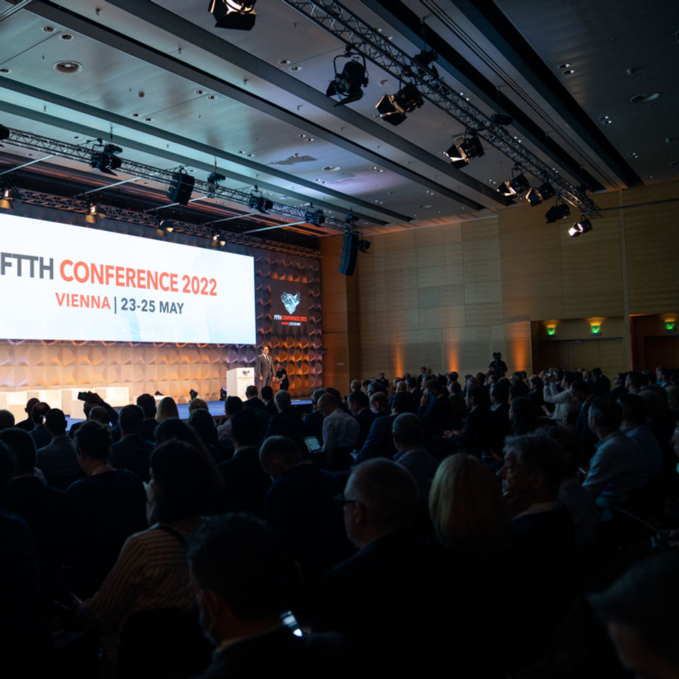 Large conference hall with a professionally lit stage featuring a screen reading “FTTH Conference 2022 – Vienna | 23–25 May” and a modern logo. A full audience attentively follows the presentation, creating a focused, high-end event atmosphere.