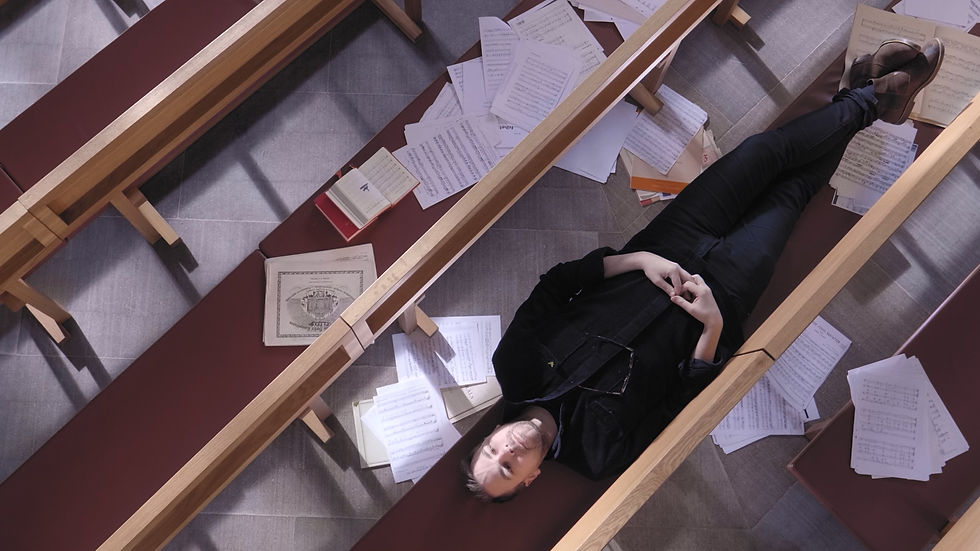 A man lays down on a church bench, seen from right above him. There’s hundreds of pages of sheet music strewn about around him, on other benches and on the floor.