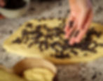 A hand sprinkles chocolate chips on vegan colomba dough on a speckled countertop. A bowl of more chips is beside. The mood is calm and focused.