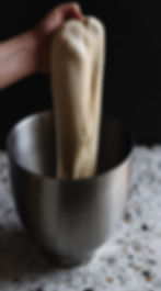 A hand stretches dough above a metal bowl on a speckled countertop. The background is dark, highlighting the dough's texture.