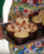 Woman in colourful Italian apron holds a tray of almond-topped vegan colomba with sugar crystals ready for the oven. Bright quilt pattern and green sleeve visible.
