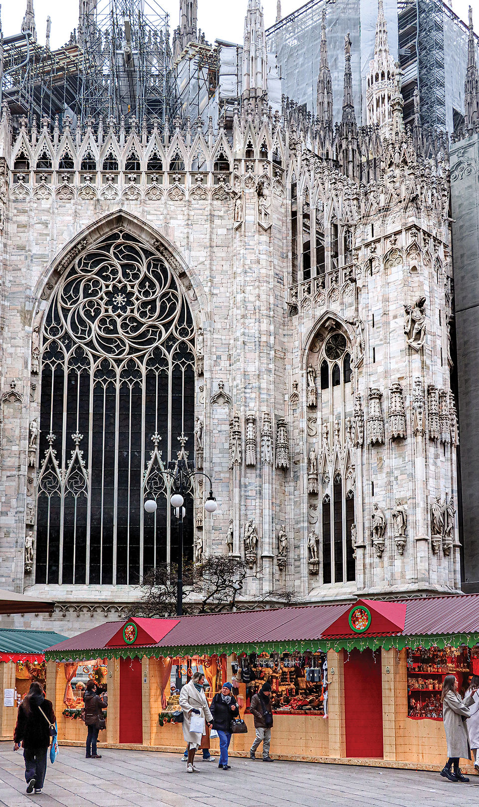 People walk past festive market stalls in front of Milan's Duomo. Stalls display colorful goods against a backdrop of intricate stonework.