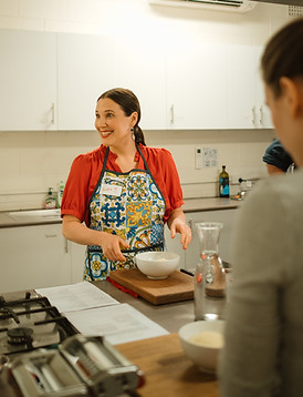 a smiling female cooking class teacher dressed in red with an Italian styled apron is leading a class
