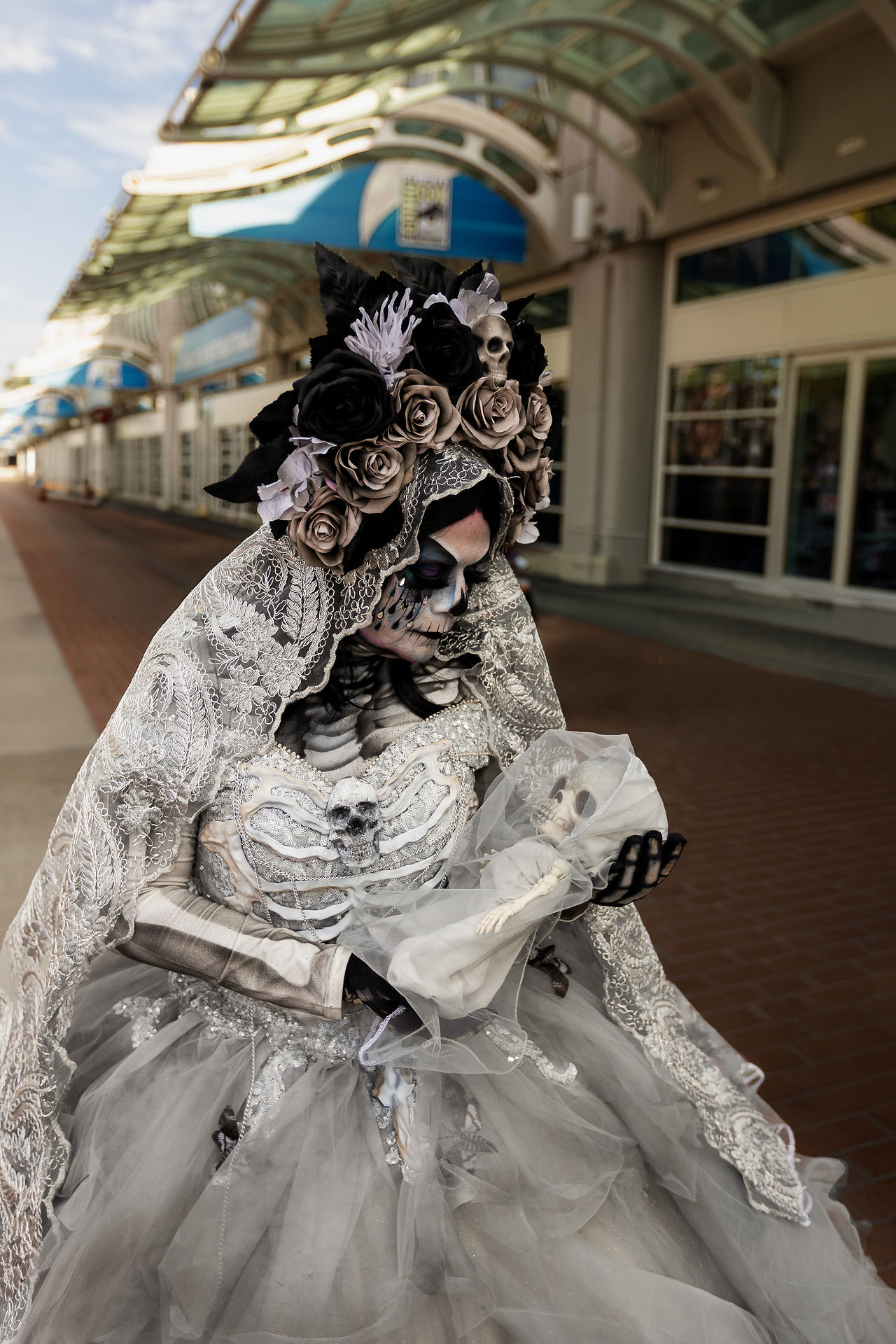 @lamuertemaria dressed as La Llorona during San Diego Comic-Con 2026, wearing skeletal makeup, an elaborate floral crown, and a lace bridal gown while cradling a skeletal infant prop outside the San Diego Convention Center.