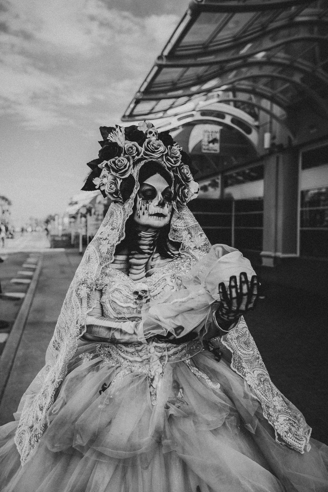 Black and white portrait of @lamuertemaria dressed as La Llorona at San Diego Comic-Con 2026, wearing skeletal makeup, a floral crown, and a lace bridal gown while holding a skeletal infant prop outside the San Diego Convention Center.