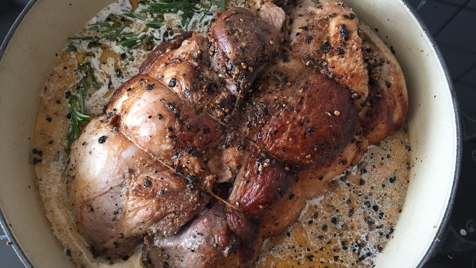 Trussed and seared pork with whole black peppercorns and rosemary braising in liquid in a large pot. Juicy and browned with herbs visible, creating a warm and savoury atmosphere. The setting is a stove top.