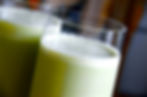 Close-up of two glasses with frothy green Matcha and banana smoothie against a blurred background, creating a fresh and inviting scene.