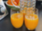 Two glasses of orange juice and a textured glass jug on a black surface. Bowl of oranges and lemons in the background. Bright, fresh mood.