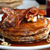 Stack of pancakes topped with crispy bacon, walnuts, and syrup on a white plate. A fork and a cup in the background. Warm, appetizing mood.