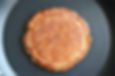 Golden-brown pancake cooking in a black frying pan, showing a crispy texture. No text visible. Warm and inviting mood.