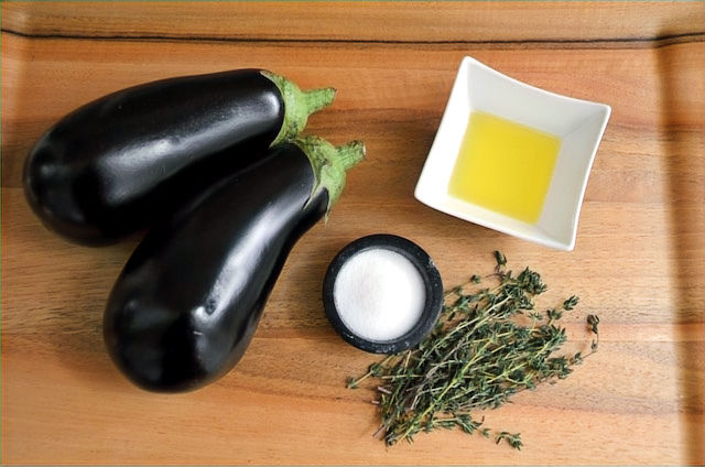 Two eggplants, a dish of olive oil, a bowl of salt, and sprigs of thyme on a wooden surface. Background is simple and natural.
