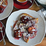 French toast topped with roasted plums, almonds, and powdered sugar, served with espresso on a wooden table. Red plates add a vibrant touch.