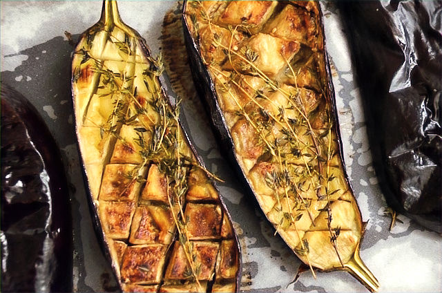Roasted eggplants on a baking sheet with a diamond pattern, garnished with thyme sprigs. Golden-brown texture, rustic feel.